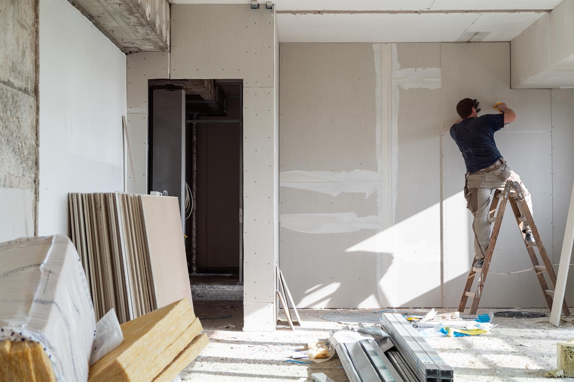 Trabajador instala paneles de yeso en interior en construcción, con escalera, herramientas y paredes sin terminar.