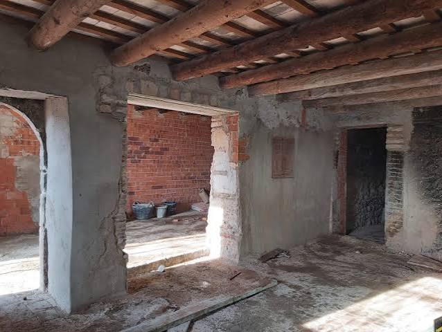 Interior en obra con paredes de ladrillo y hormigón, vigas de madera y escombros en el suelo.